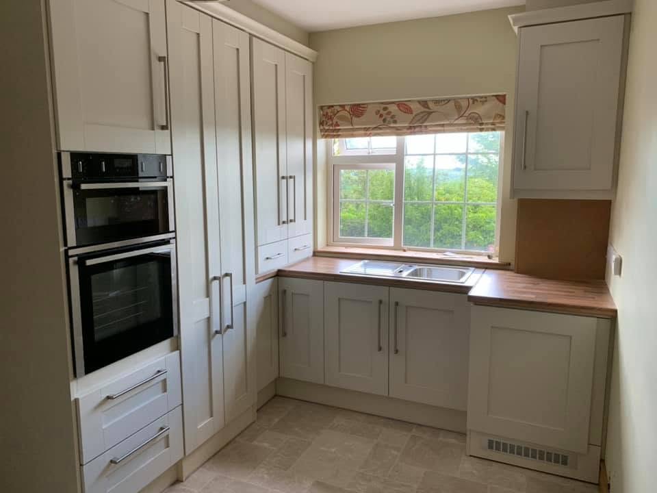 A cream painted shaker kitchen with a timber worktop, built-in ovens and a window over the sink.